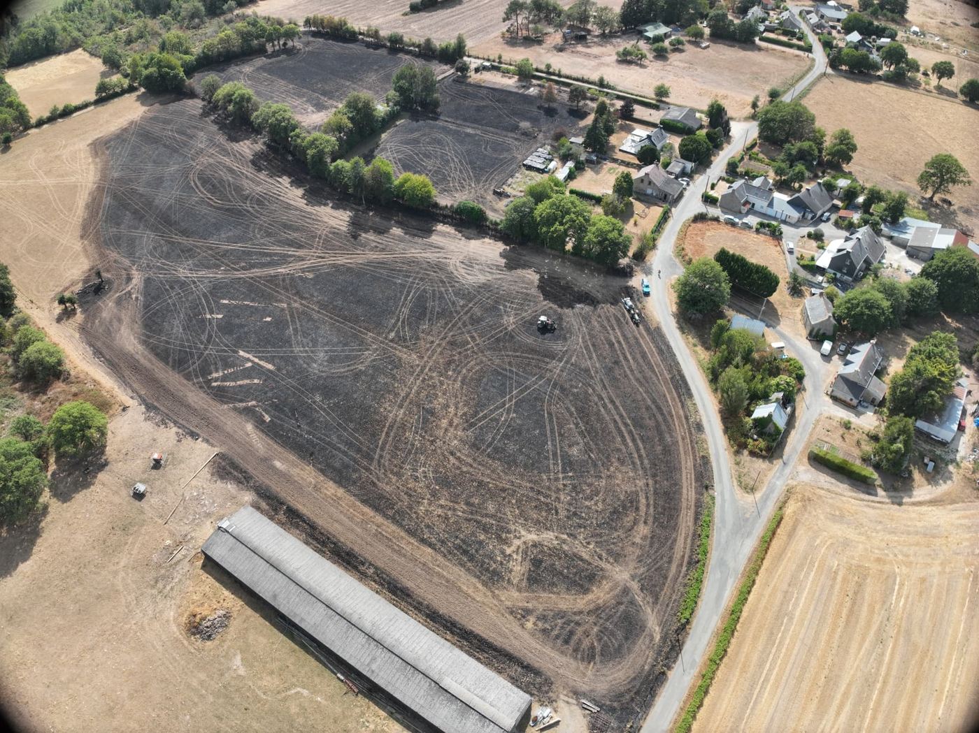 Vue aérienne par drone d'une parcelle agricole entièrement noircie par un incendie dans le Morbihan, avec un hangar agricole au premier plan et des maisons en lisière